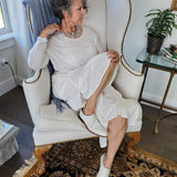 A middle aged woman sitting in an arm chair wearing a Carolily Finery statement necklace made from silver chain, European crystal pearls and European crystal