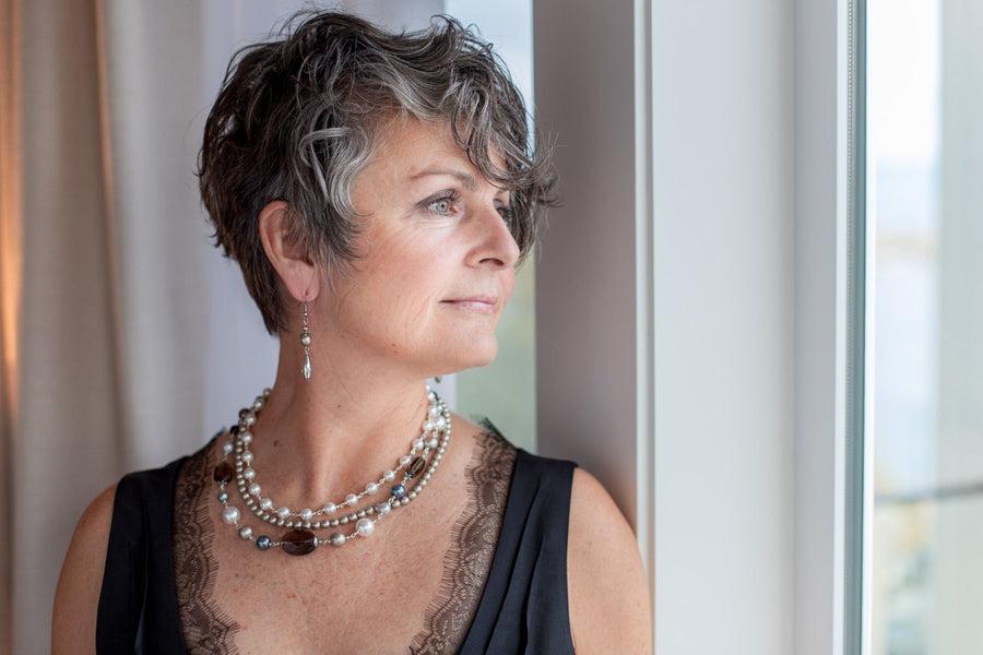 A woman looking out a window wearing a Carolily Finery statement necklace made from European crystal pearls in white, grey and platinum, and gemstones