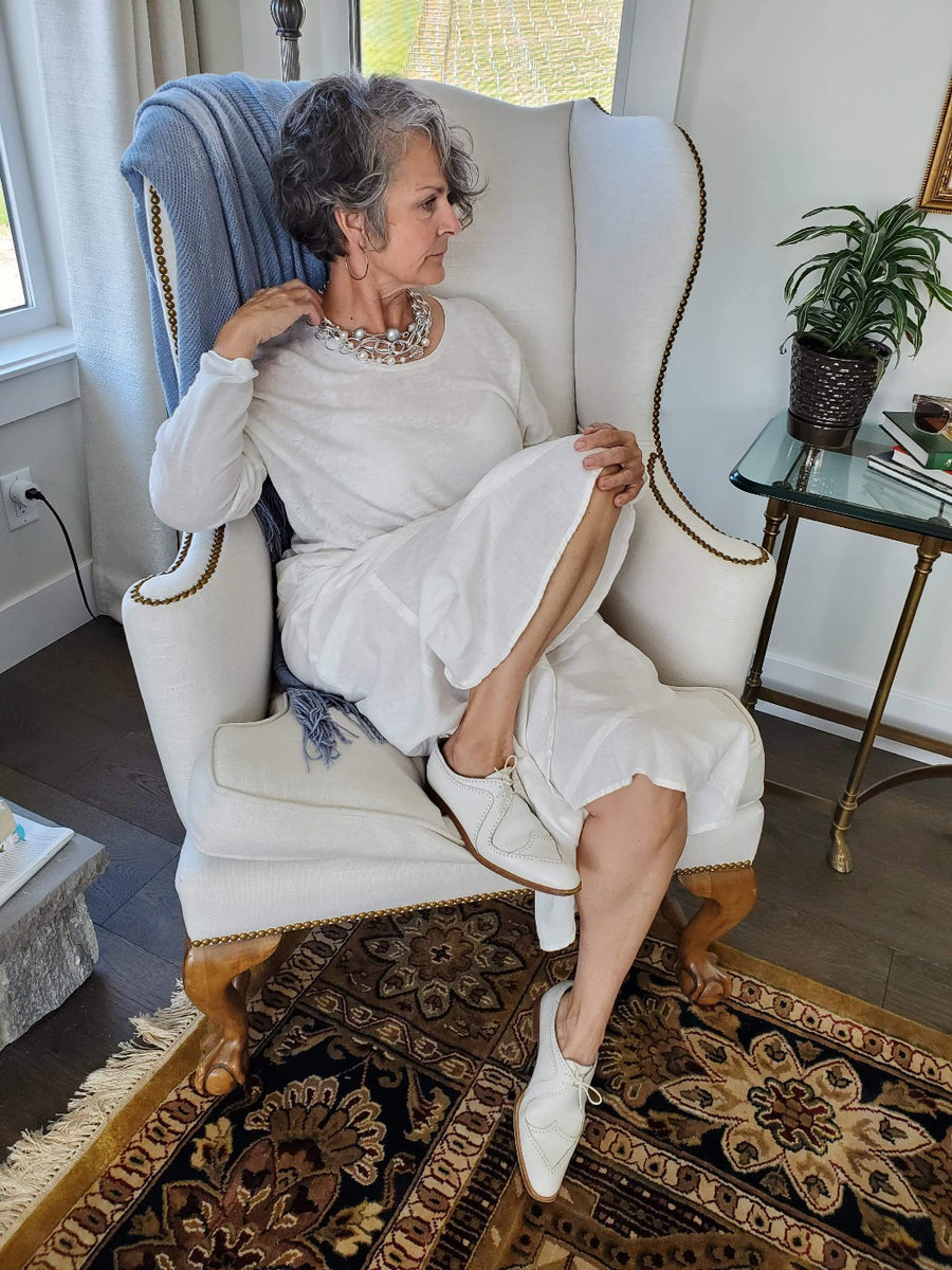 A middle aged woman sitting in an arm chair wearing a Carolily Finery statement necklace made from silver chain, European crystal pearls and European crystal